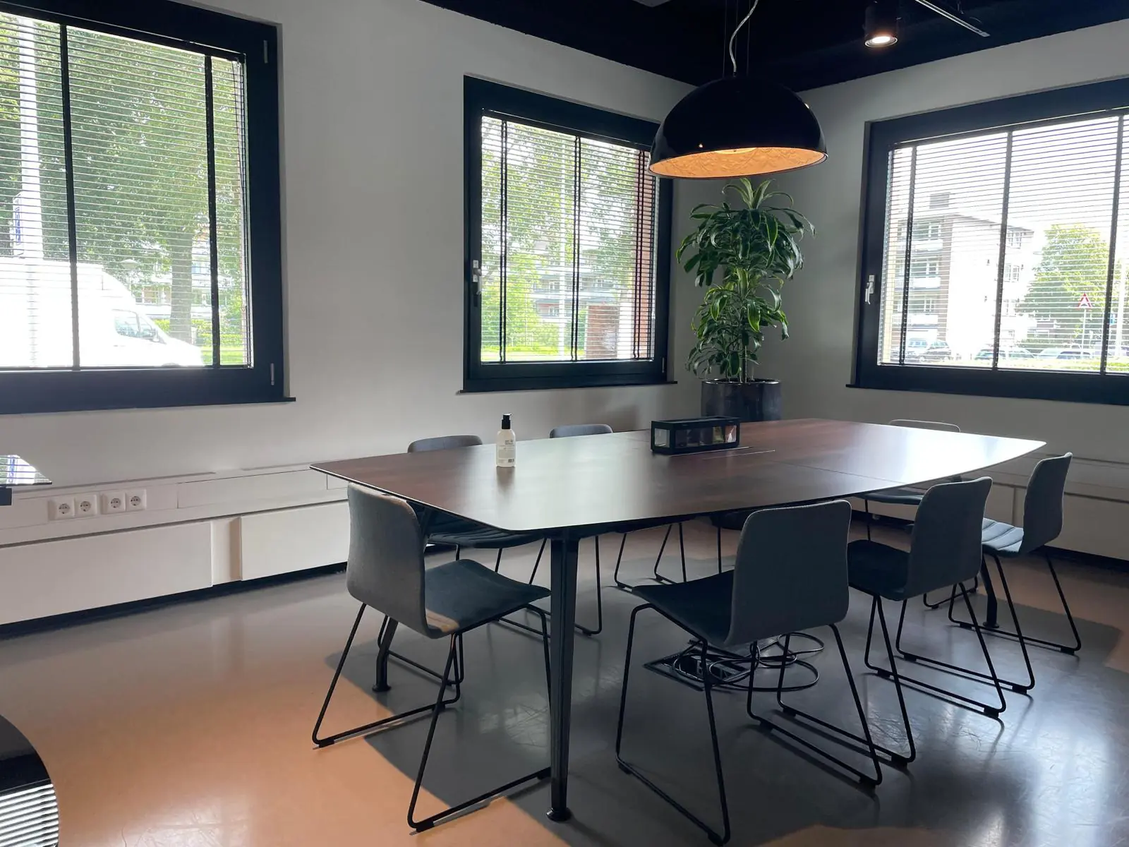 Vergaderruimte aan de De Cuserstraat met grote tafel, zes stoelen, kamerplant en hanglamp bij drie ramen met jaloezieën.